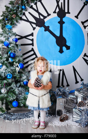 Girl holding un cône de pin. L'horloge dans l'arrière-plan afficher cinq minutes avant minuit. La veille de Noël. Nouvelle Année. Maison de vacances et de plaisir. Banque D'Images