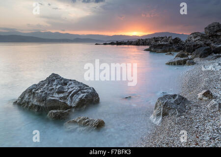 Les cailloux et les rochers sur la plage de Bataria, Kassiopi, Corfou sont éclairés avec la forte lumière orange de lever du soleil. Banque D'Images