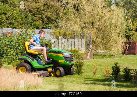 Un garçon de 15 ans de tondre la pelouse sur un John Deere ride sur la faucheuse au Royaume-Uni Banque D'Images