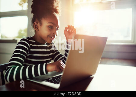 Petite fille assise ethnique à la maison à une table à travailler sur ses devoirs sur un ordinateur portable la lecture de l'écran avec un air pensif Banque D'Images