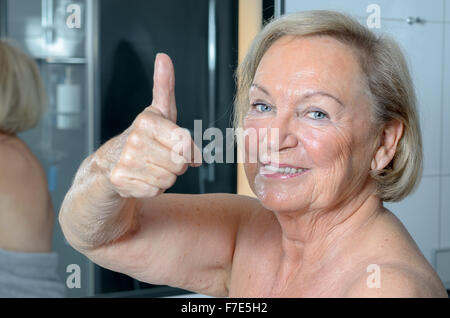 Attractive blonde woman standing dans une serviette dans une salle de bains tout en donnant un geste vers le pouce, Close up portrait of il Banque D'Images