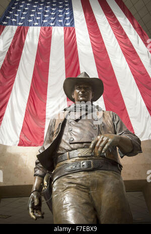 Le 27 novembre 2015 - Santa Ana, Californie, États-Unis - une plus grande que la vie statue en bronze de John Wayne est sur l'affichage à l'aéroport international John Wayne. Wayne, un acteur célèbre pour ses rôles de cow-boy, le vécu dans les environs de Newport Beach. La statue de bronze de Wayne est encadrée par un drapeau américain fond à partir derrière illustrant une image de patriotisme pour une génération qui a grandi sur ses films représentant l'individualisme farouche et emblématiques de l'époque beaucoup aimé dans l'histoire américaine et de la culture. Wayne est représenté, comme il l'a été dans la plupart de ses films de l'ouest, comme individualiste robuste, hea Banque D'Images