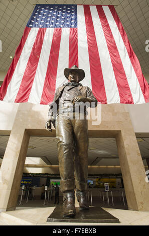 Le 27 novembre 2015 - Santa Ana, Californie, États-Unis - une plus grande que la vie statue en bronze de John Wayne est sur l'affichage à l'aéroport international John Wayne. Wayne, un acteur célèbre pour ses rôles de cow-boy, le vécu dans les environs de Newport Beach. La statue de bronze de Wayne est encadrée par un drapeau américain fond à partir derrière illustrant une image de patriotisme pour une génération qui a grandi sur ses films représentant l'individualisme farouche et emblématiques de l'époque beaucoup aimé dans l'histoire américaine et de la culture. Wayne est représenté, comme il l'a été dans la plupart de ses films de l'ouest, comme individualiste robuste, hea Banque D'Images