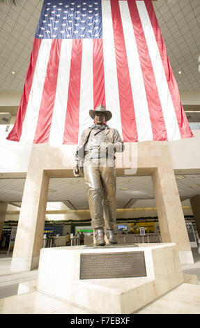 Le 27 novembre 2015 - Santa Ana, Californie, États-Unis - une plus grande que la vie statue en bronze de John Wayne est sur l'affichage à l'aéroport international John Wayne. Wayne, un acteur célèbre pour ses rôles de cow-boy, le vécu dans les environs de Newport Beach. La statue de bronze de Wayne est encadrée par un drapeau américain fond à partir derrière illustrant une image de patriotisme pour une génération qui a grandi sur ses films représentant l'individualisme farouche et emblématiques de l'époque beaucoup aimé dans l'histoire américaine et de la culture. Wayne est représenté, comme il l'a été dans la plupart de ses films de l'ouest, comme individualiste robuste, hea Banque D'Images