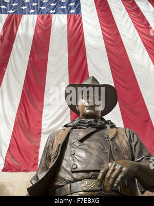 Le 27 novembre 2015 - Santa Ana, Californie, États-Unis - une plus grande que la vie statue en bronze de John Wayne est sur l'affichage à l'aéroport international John Wayne. Wayne, un acteur célèbre pour ses rôles de cow-boy, le vécu dans les environs de Newport Beach. La statue de bronze de Wayne est encadrée par un drapeau américain fond à partir derrière illustrant une image de patriotisme pour une génération qui a grandi sur ses films représentant l'individualisme farouche et emblématiques de l'époque beaucoup aimé dans l'histoire américaine et de la culture. Wayne est représenté, comme il l'a été dans la plupart de ses films de l'ouest, comme individualiste robuste, hea Banque D'Images