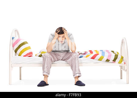 Jeune mec en pyjama assis sur un lit et souffrant d'insomnie isolé sur fond blanc Banque D'Images