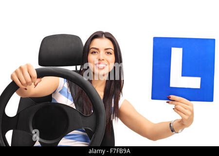 Jeune femme tenant un volant et une L-sign assis sur un siège de voiture isolé sur fond blanc Banque D'Images