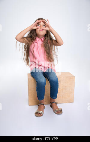 Portrait d'une petite fille assise sur boîte en bois et à la recherche à travers les doigts isolé sur fond blanc Banque D'Images