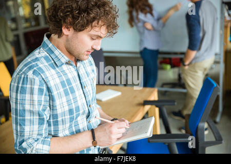 Jeune homme confiant réfléchie bouclés en chemise à carreaux bleu des notes dans l'ordinateur portable et la création de projet avec des collègues Banque D'Images