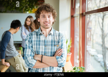 Jeune mâle curly positifs attrayants en chemise à carreaux posant dans office avec les mains pliées, debout près de la fenêtre Banque D'Images