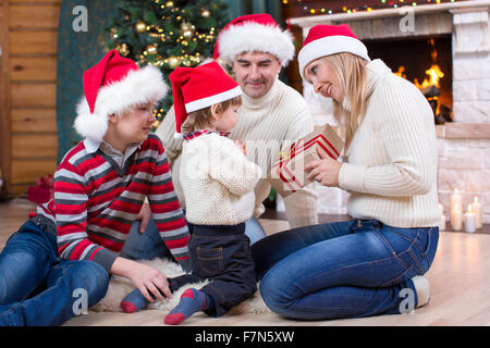 Famille heureuse en bonnets rouges avec don assis à arbre de Noël près de cheminée Banque D'Images