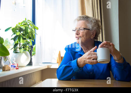 Grand-mère de boire du café et à l'extérieur de sa fenêtre dans la maison pour les personnes âgées Banque D'Images