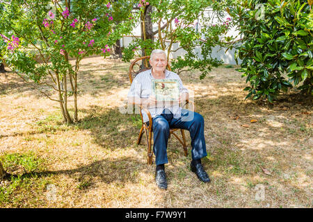 Personnes âgées L'octogénaire homme souriant et montrant old vintage photo dans un cadre tout en restant assis sur le fauteuil en osier dans le jardin Banque D'Images