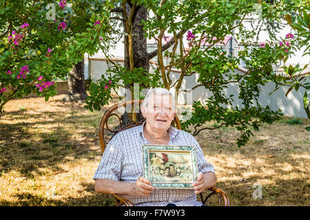 Personnes âgées L'octogénaire homme souriant et montrant old vintage photo dans un cadre tout en restant assis sur le fauteuil en osier dans le jardin Banque D'Images