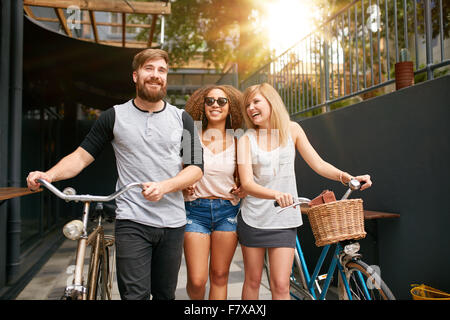 Trois jeunes gens marchant dans la rue avec leurs bicyclettes et d'avoir du plaisir. Les amis masculins et féminins avec leur vélo sur city s Banque D'Images