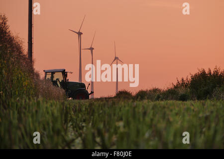 Un tracteur traversant le décor d'un domaine de pailles de blé fauchés et quelques éoliennes dans l'arrière-plan sous un ciel rouge Banque D'Images