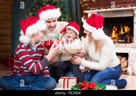 Famille heureuse en bonnets rouges avec des cadeaux assis à arbre de Noël près de cheminée Banque D'Images