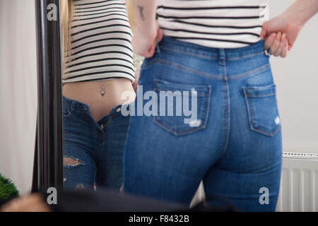 Une femme à la maison en tirant sur des jeans serrés devant un miroir de chambre à coucher. Banque D'Images