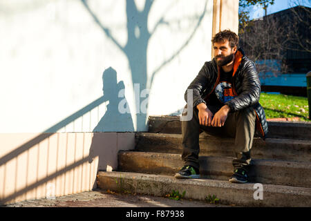 Barbu brun jeune garçon assis sur des escaliers street bestido avec plumes gris et orange , un T-shirt noir, un pantalon vert et sneake Banque D'Images