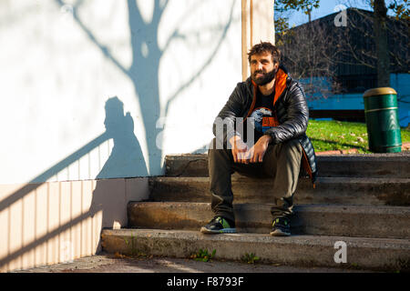 Barbu brun jeune garçon assis sur des escaliers street bestido avec plumes gris et orange , un T-shirt noir, un pantalon vert et sneake Banque D'Images