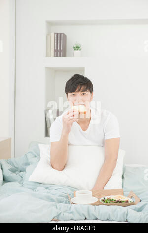 Homme assis dans le lit de mordre une tranche de pain avec le plateau du petit déjeuner Banque D'Images