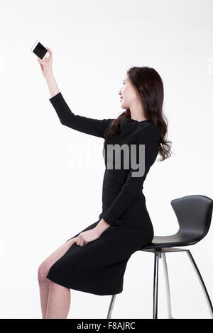 Jeune femme en robe noire assise sur une chaise et prendre une photo avec un smartphone Banque D'Images