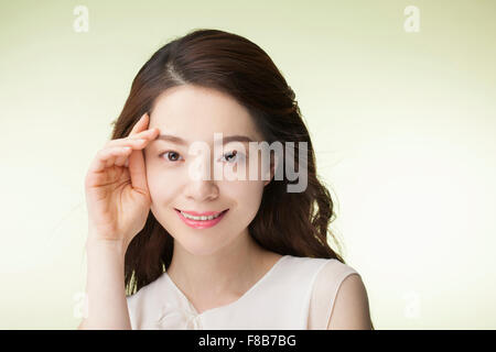 Femme coréenne avec la longue chevelure ondulée en blanc T-shirt mettant sa main sur son visage à côté de ses yeux et souriant Banque D'Images