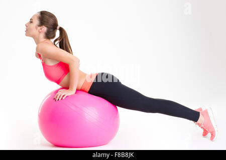 L'accent séduisant jeune athlète en survêtement faisant des exercices d'étirement sur fitball rose sur fond blanc isolé Banque D'Images