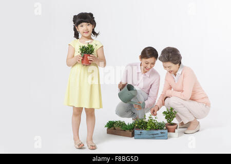 Petite-fille tenant une plante et mère et grand-mère d'arroser d'autres plantes sur le sol derrière la petite-fille Banque D'Images