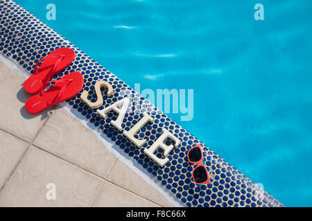 High angle de vente caractères placés sur la margelle de piscine en entre une paire de tongs et lunettes de soleil Banque D'Images