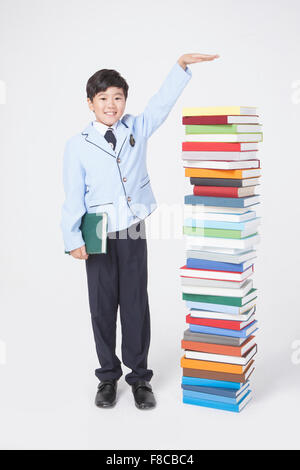 Petit garçon de l'école élémentaire en uniforme tenant un livre et debout à côté d'une pile de livres avec un geste de la main signifiant Banque D'Images