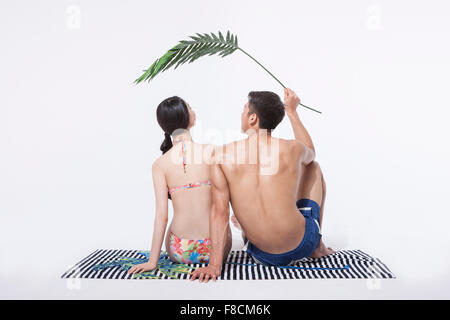 Retour apparition d'un couple dans les vêtements de plage assis sur une serviette avec les feuilles des plantes tenues par un homme au-dessus de leurs têtes Banque D'Images