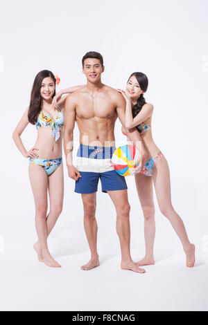 Deux femmes en bikini s'appuyant sur un homme musclé en natation pantalon dans entre eux tenant un ballon de plage tous les comités permanents et fixant Banque D'Images