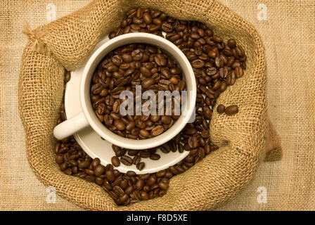 Tasse et soucoupe blanc rempli de grains de café assis dans un sac de toile rempli de grains de café sur une toile de fond. Banque D'Images