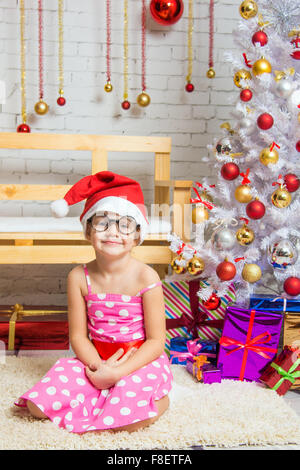 Girl in the red hat et le funny lunettes rondes est assis sur un tapis dans la chambre à l'intérieur du Nouvel An Banque D'Images