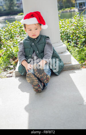 Mélancolie Mixed Race Boy Sitting At The Park Wearing Christmas Santa Hat. Banque D'Images