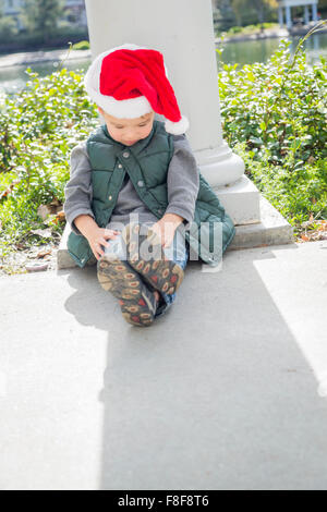 Mélancolie Mixed Race Boy Sitting At The Park Wearing Christmas Santa Hat. Banque D'Images