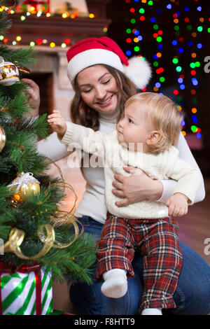 Mère en rouge santa hat la regarde d'un peu petit garçon souriant et près des arbres de Noël Banque D'Images