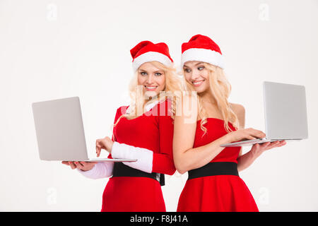 Belle blonde gaie sœurs jumeaux en rouge santa claus les vêtements et les chapeaux à l'aide de deux ordinateurs portables sur fond blanc Banque D'Images