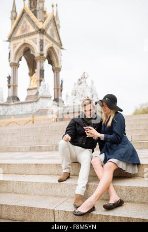 Young couple sitting on steps reading textes smartphone, Albert Memorial, London, England, UK Banque D'Images