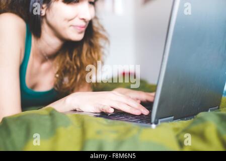 Jeune femme couchée sur le lit à l'aide avant de l'ordinateur portable en souriant, differential focus Banque D'Images