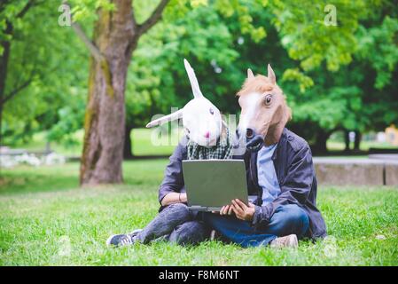 Young couple sitting on grass portant des masques de lapin et l'ordinateur portable costume holding Banque D'Images