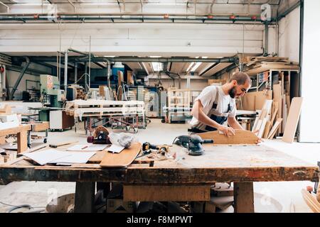 Jeune homme en atelier de menuiserie à la recherche vers le bas de la planche à roulettes de ponçage Banque D'Images