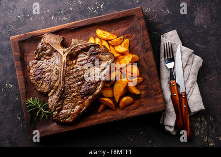 Rare moyen T-bone steak grillé avec les quartiers de pommes de terre sur le service à bord bloc sur fond sombre Banque D'Images