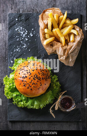 Des hamburger avec graines de sésame noir et de frites de pommes de terre dans le papier dorsal, servi avec sauce ketchup dans verre Banque D'Images