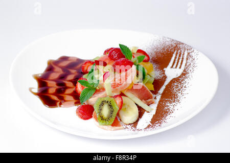 Close-up de la salade de fruits, décoré d'leaces la menthe, la cannelle en poudre et la sauce brune, sur la plaque ovale, blanc sur fond blanc Banque D'Images