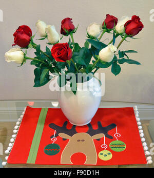 Roses rouges et blanches dans un vase en porcelaine blanche reste sur un tapis de table sur le thème de Noël avec un design contemporain représentant un renne stylisé avec ornements de vacances suspendu à ses bois. Banque D'Images