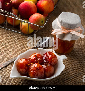 Pommes cerises au sirop couvert sur blanc soucoupe, cuillère, pot de confiture faite maison et des pommes en fil métallique panier sur une table cove Banque D'Images