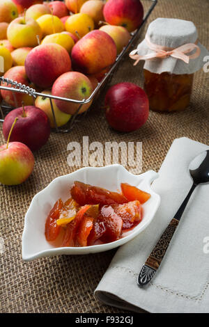 Des tranches de pomme confiture sur la soucoupe blanc, cuillère, pot de confiture, de linge de maison serviette et pommes en fil métallique panier sur une toile cov Banque D'Images