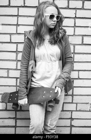 Adolescente en jeans et lunettes détient plus de skateboard urban mur de briques, l'arrière-plan photo monochrome Banque D'Images